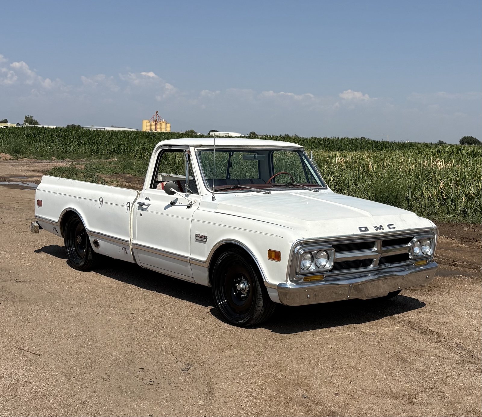 White late-1960s GMC pickup on black steel wheels beside a cornfield